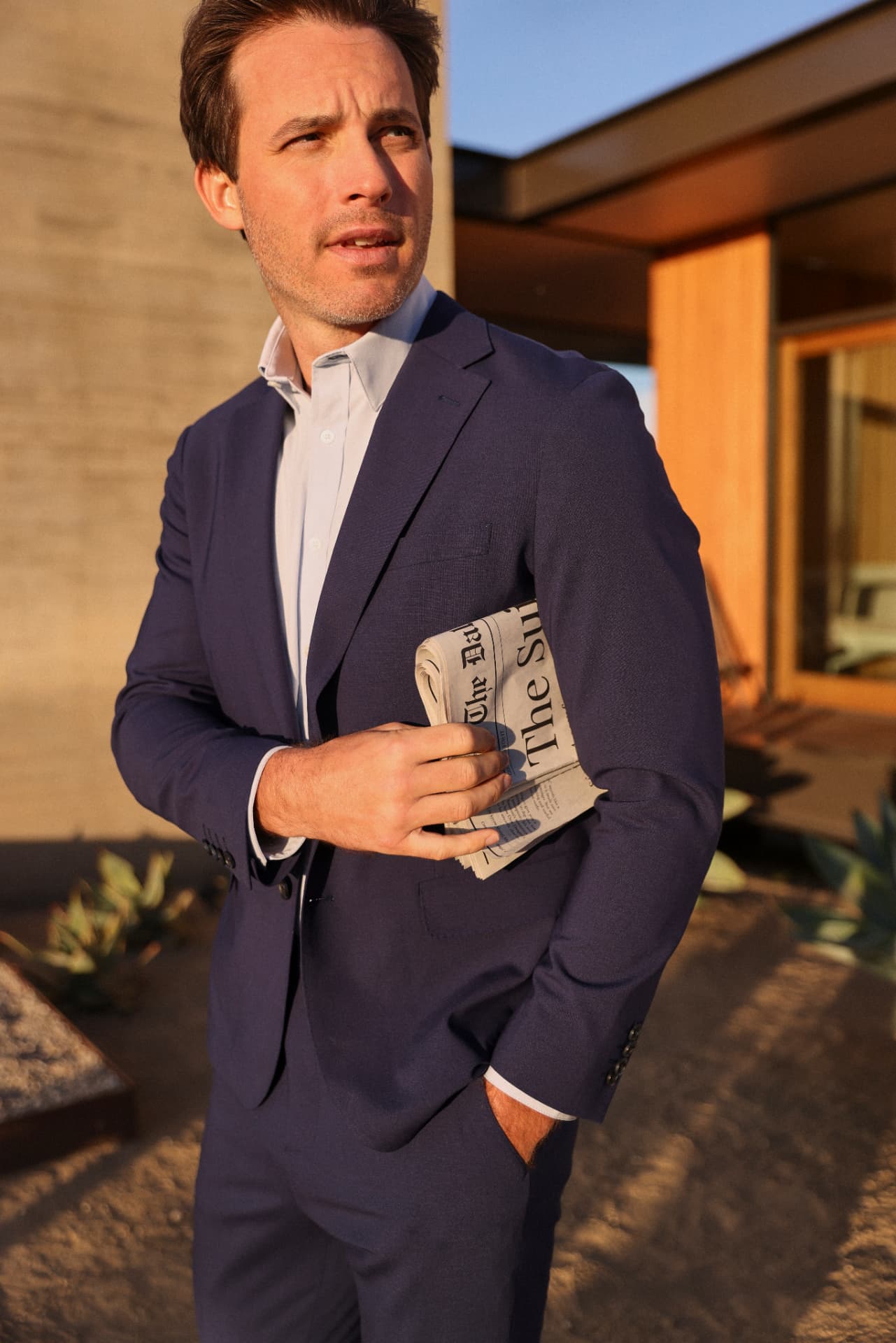 Man wearing a navy suit and pale blue shirt, jacket buttoned, in evening light