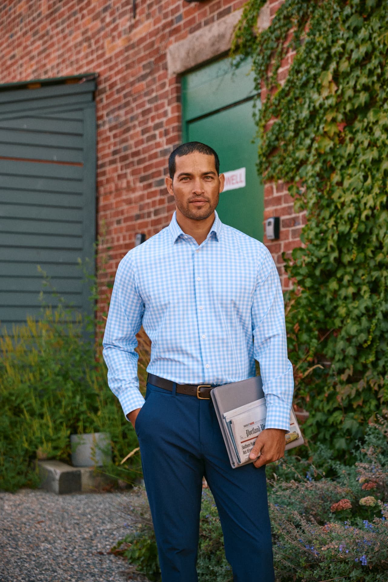 Man in a light blue checked button-down shirt and navy trousers outside a brick building