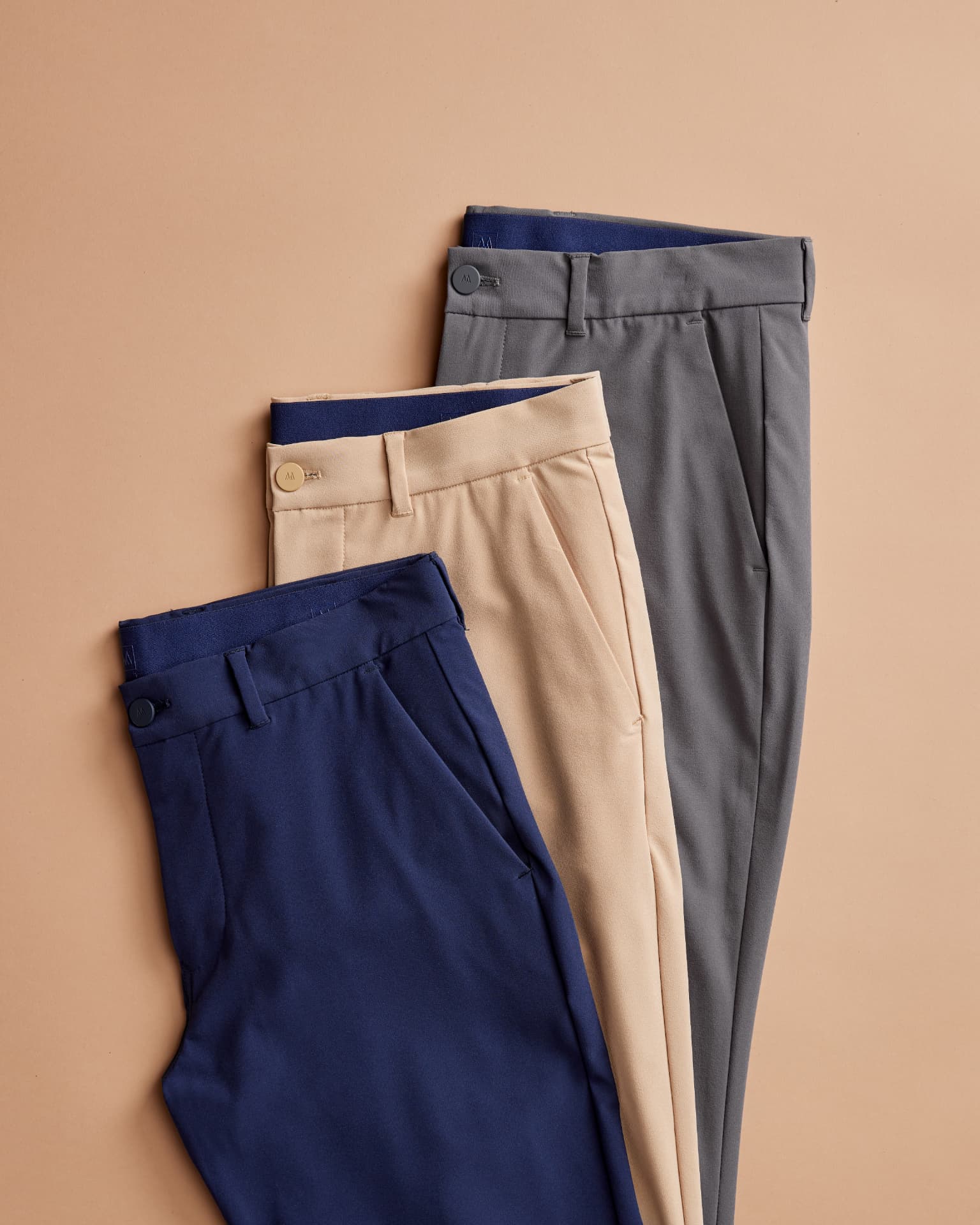 Three pairs of folded trousers in navy, tan, and grey laid flat