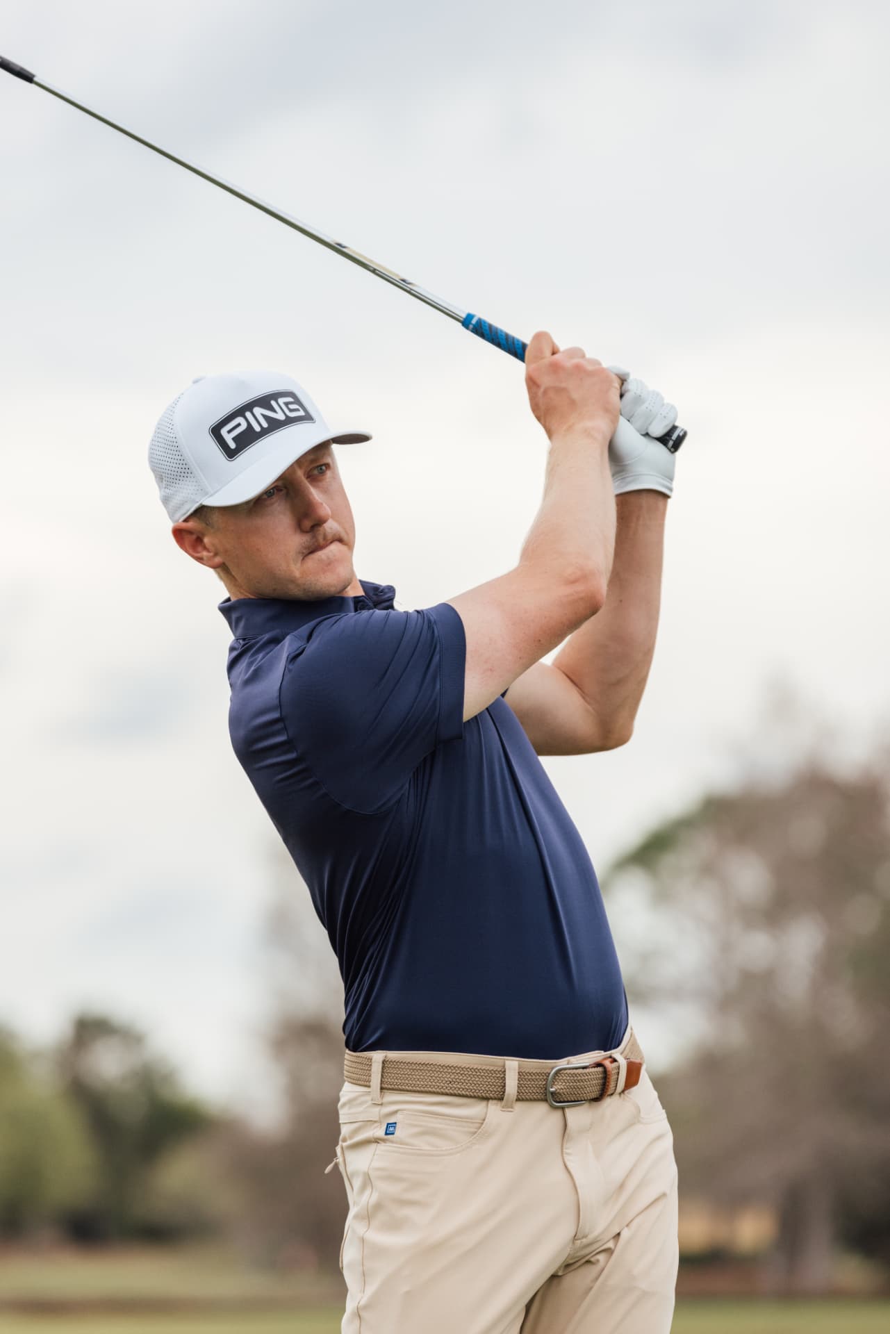 Man in a navy performance polo, tan trousers, and a white cap finishing a golf swing