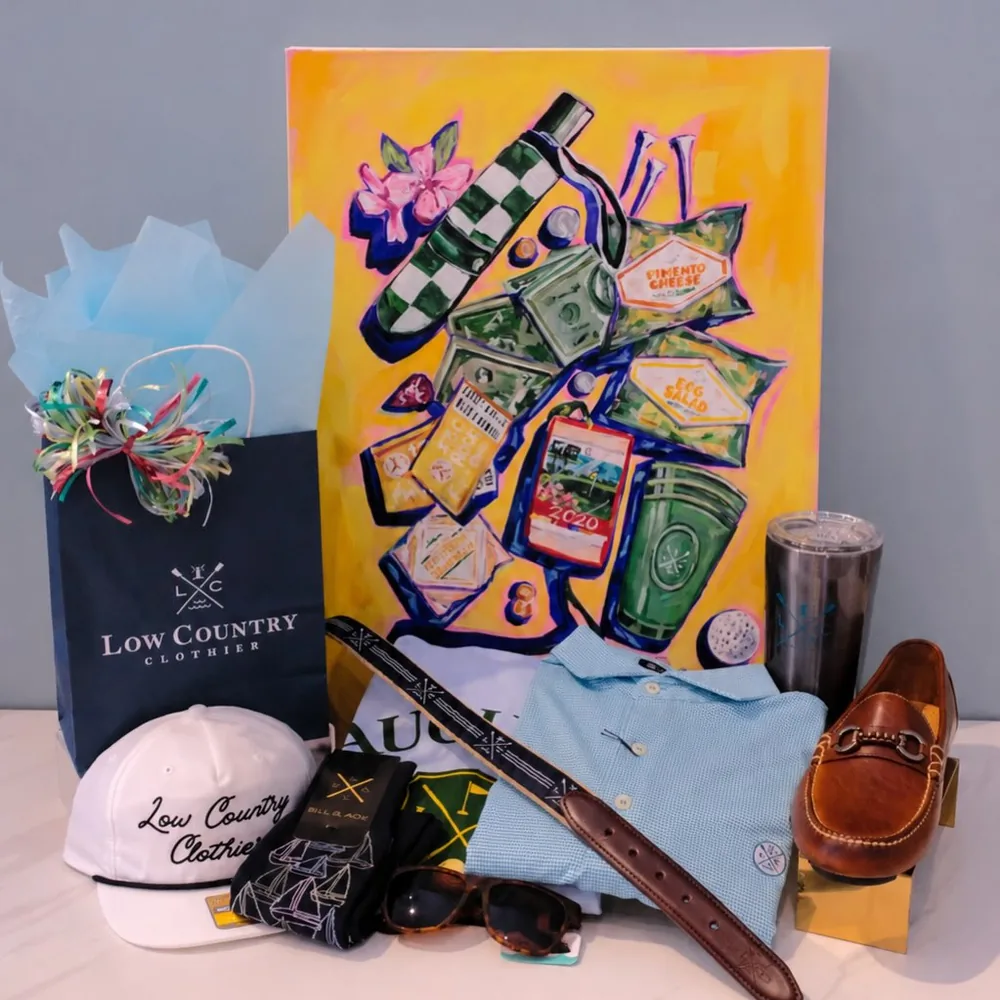 Low Country Clothier tote bag and cap styled with a blue polo, woven belt, sunglasses and leather loafers in front of a colorful painting