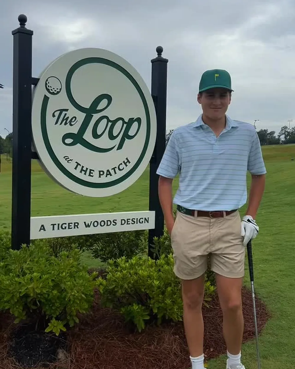 Man in a striped polo, khaki shorts and cap standing with a golf club beside The Loop at The Patch sign